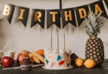 A colorful birthday cake with fruits and festive decorations on a table indoors.