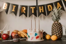 A colorful birthday cake with fruits and festive decorations on a table indoors.