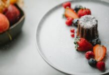 From above of palatable homemade pie sprinkled with icing sugar placed on round plate on table near blurred bowl in kitchen