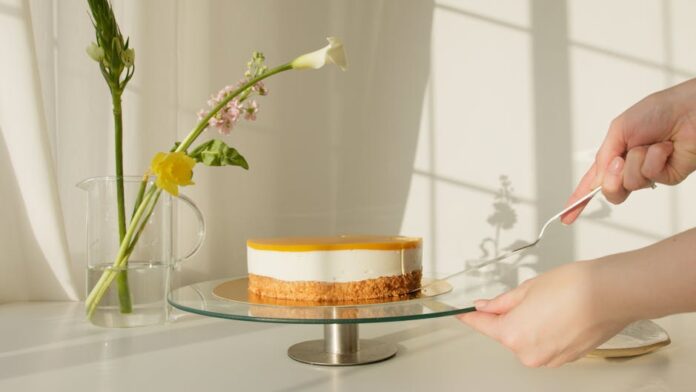 Bolo de Iogurte com Limão e Mel: O Sabor que Surpreende! Elegant lemon cheesecake on a stand surrounded by natural flowers. Perfect for celebrations or culinary projects.