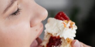 Crop unrecognizable female with closed eyes biting tasty sweet cupcake with with strawberry jam filing and decorated with whipped cream and berries
