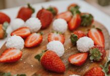 Sobremesa fácil sem forno que vai surpreender Appetizing strawberries and coconut balls arranged on a wooden board, perfect for dessert lovers.