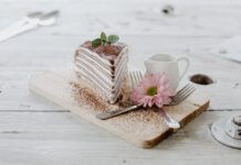 Torta de chocolate com recheio cremoso irresistível From above of appetizing piece of cake decorated chocolate powder and mint leaves served near ceramic creamer and forks with light pink chrysanthemum on top placed on wooden board