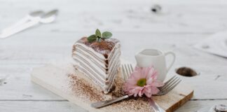 From above of appetizing piece of cake decorated chocolate powder and mint leaves served near ceramic creamer and forks with light pink chrysanthemum on top placed on wooden board
