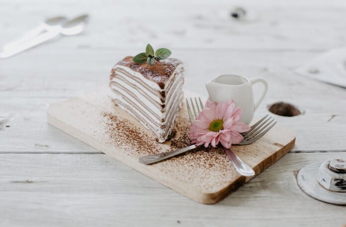 Receita de torta de chocolate com recheio cremoso irresistível From above of appetizing piece of cake decorated chocolate powder and mint leaves served near ceramic creamer and forks with light pink chrysanthemum on top placed on wooden board