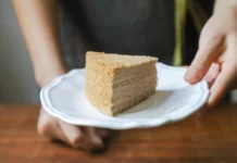 Bolo Caseiro económico e fofinho: O segredo da maciez Hand holding a piece of homemade cake on a white plate, offering it as a treat.