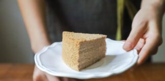 Hand holding a piece of homemade cake on a white plate, offering it as a treat.