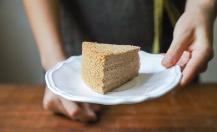 Bolo Caseiro Económico e Fofinho: O Segredo da Maciez! Hand holding a piece of homemade cake on a white plate, offering it as a treat.