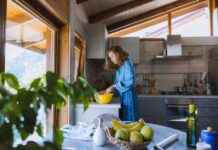 Bolo de Banana com Rum A woman with curly hair prepares a meal in a sunlit kitchen, surrounded by fresh fruits.