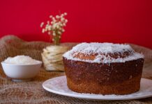 Bolo de coco húmido caseiro: Descubra o segredo da receita A mouthwatering homemade coconut cake with shredded coconut topping against a vibrant red backdrop.