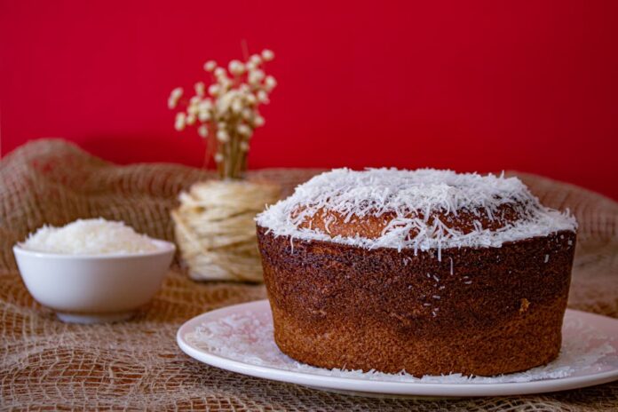 Bolo de coco húmido caseiro: descubra o segredo da receita! A mouthwatering homemade coconut cake with shredded coconut topping against a vibrant red backdrop.