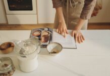 Bolo rápido para iniciantes Hands pointing at cookbook in a cozy kitchen setting with ingredients ready for baking.
