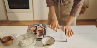 Hands pointing at cookbook in a cozy kitchen setting with ingredients ready for baking.