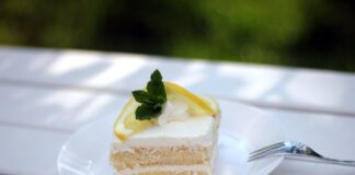 Delicious lemon cream cake slice garnished with lemon and mint on white plate.