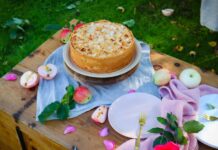 Receita de Bolo de Maçã simples para o dia a dia A rustic apple cake topped with almonds on a wooden table outdoors, accented by fresh apples and petals.