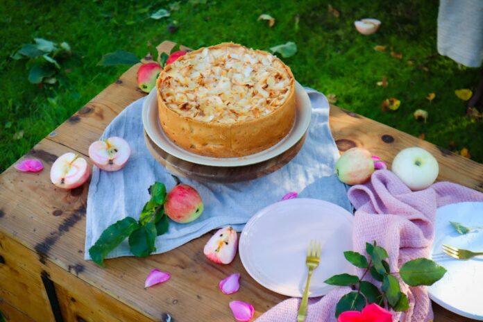 Receita de Bolo de Maçã Simples para o Dia a Dia: Descubra! A rustic apple cake topped with almonds on a wooden table outdoors, accented by fresh apples and petals.