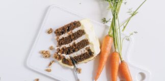 A tantalizing slice of carrot cake with frosting, walnuts, and fresh carrots on a chopping board.