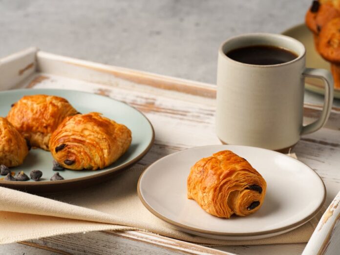 Descubra os Segredos do Croissant Doce Perfeito em Casa Enjoy a delightful breakfast with fresh croissants and a cup of coffee served on a rustic tray.