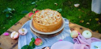 A rustic apple cake topped with almonds on a wooden table outdoors, accented by fresh apples and petals.