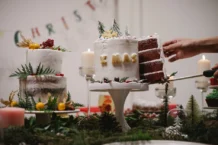 Receita de bolo sem batedeira: Fácil e surpreendente A hand cuts a slice from a beautifully decorated Christmas cake adorned with festive decor indoors.