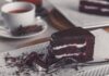 Closeup of a rich chocolate cake slice on a plate with a tea cup in the background.