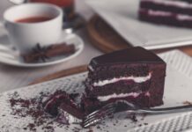 Closeup of a rich chocolate cake slice on a plate with a tea cup in the background.