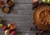 Top view of a rustic apple cake with spices and leaves on a wooden table.