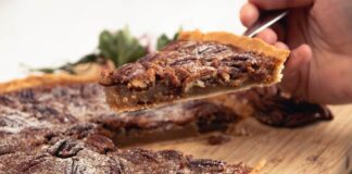 Close-up of a hand holding a sliced piece of homemade pecan pie with a sprinkle of powdered sugar.