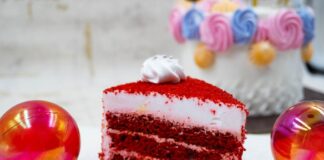 A vibrant red velvet cake slice with frosting, surrounded by festive colorful decorations.